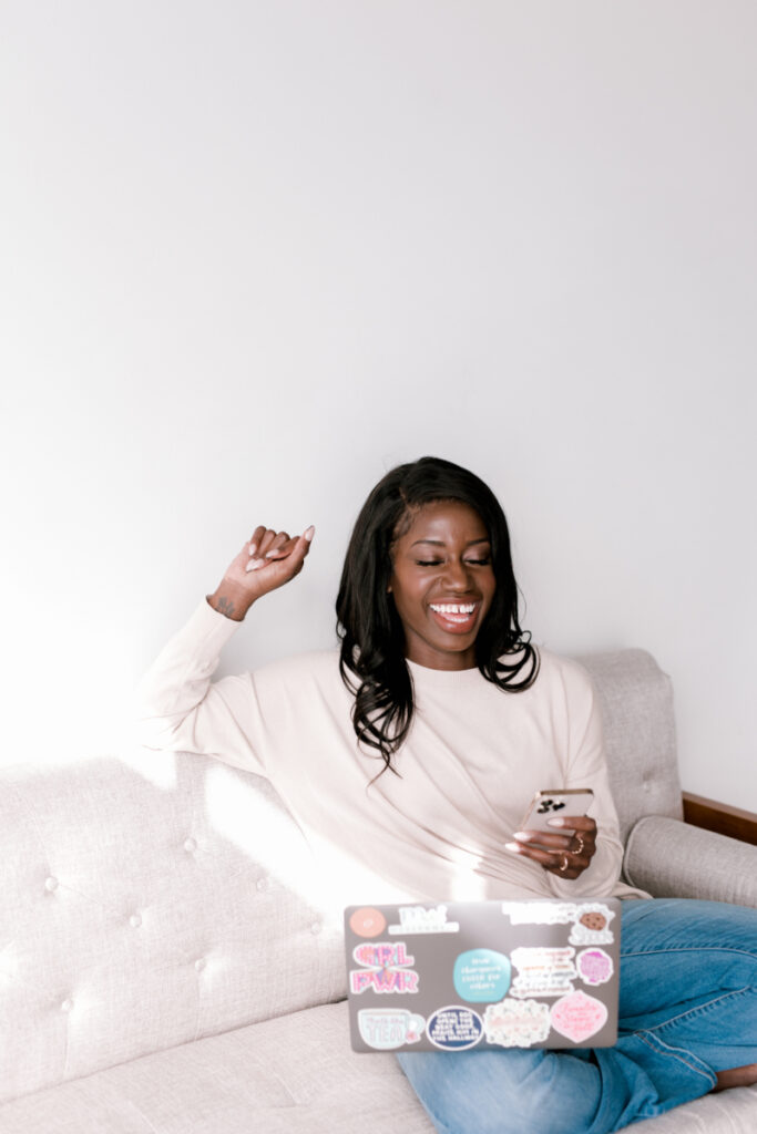 A woman laughs while sitting on a light gray sofa with a sticker-covered laptop and a smartphone. She has one arm raised in a joyful, celebratory gesture.